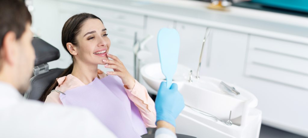 Smiling,Woman,Looking,In,Mirror,After,Dental,Procedure,In,Clinic,